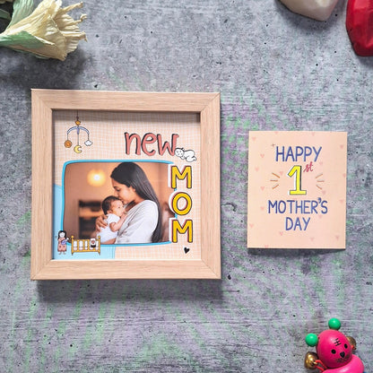 Wooden photo frame with 'New Mom' text and a small card with 'Happy 1st Mother's Day' text on a gray surface.